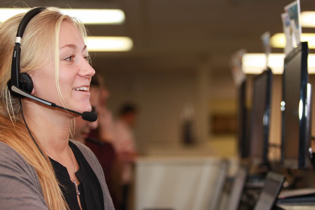 A rokrbox caller wearing a headset smiles while talking with a lead at her workstation, with other callers on shift behind her